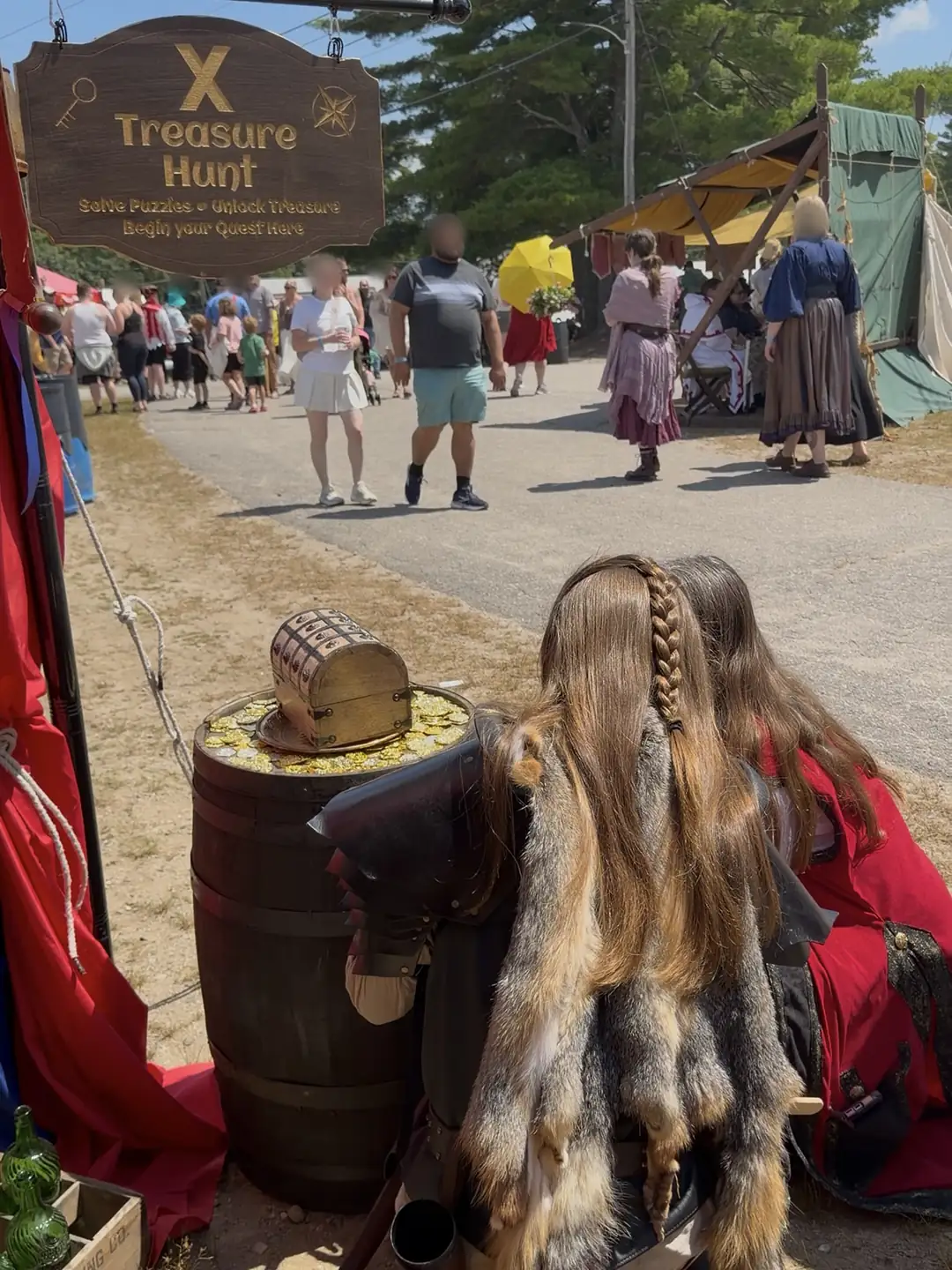 The hanging X Treasure Hunt sign over a barrel of gold coins, players in fur cloaks at the barrel and a busy faire lane behind