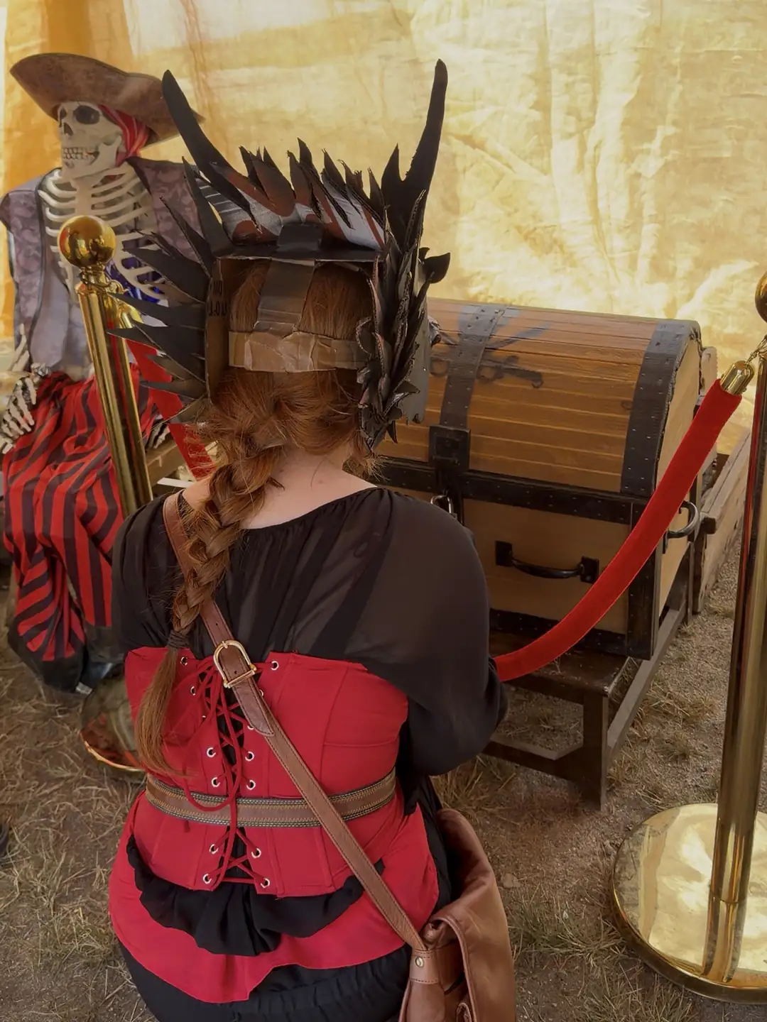A player in a feathered helm and red corset kneeling at the locked treasure chest, beside the tent's pirate skeleton
