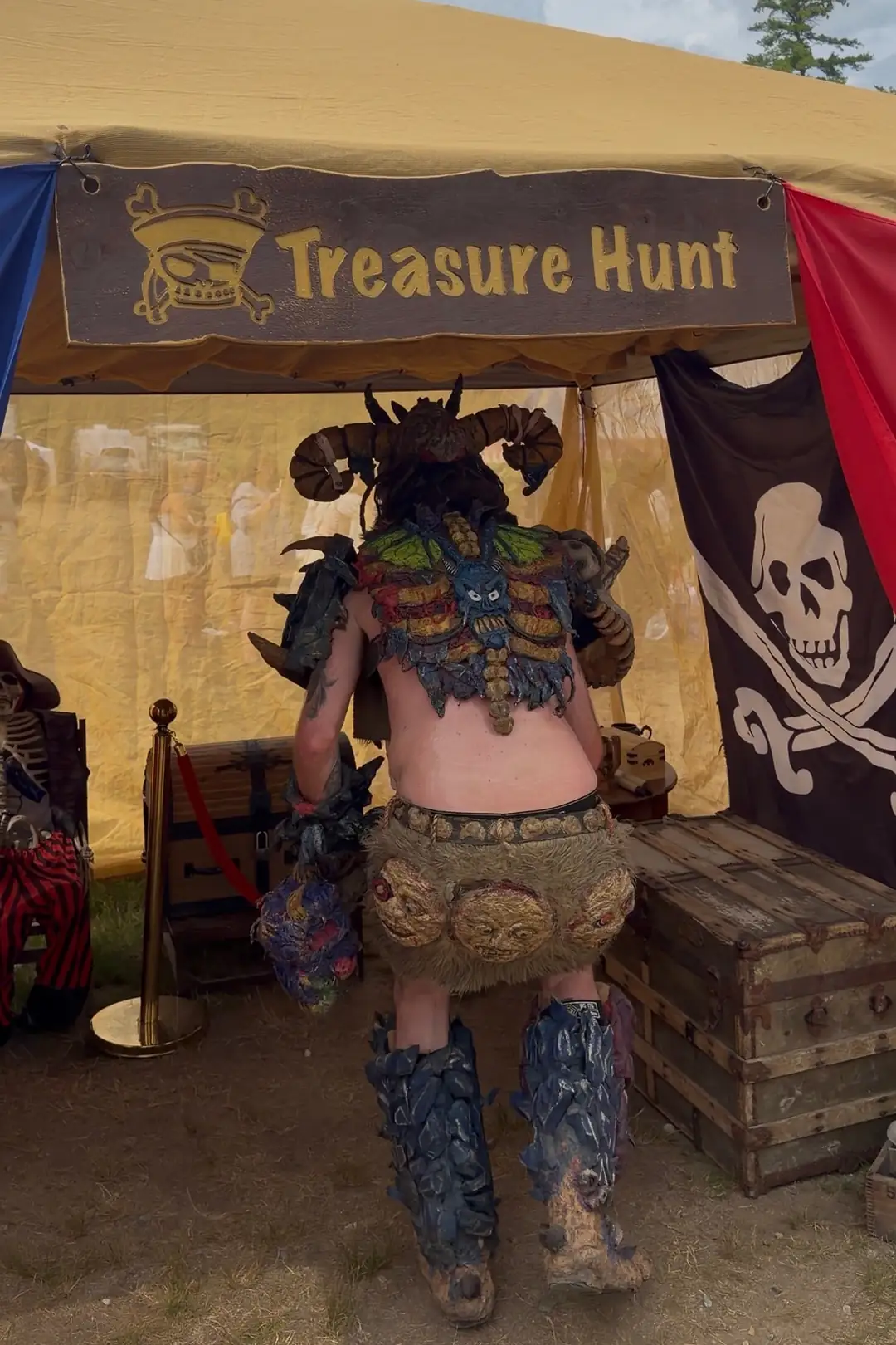 A player in a horned creature costume, seen from behind, at the X Treasure Hunt tent beside the treasure chests and the Jolly Roger