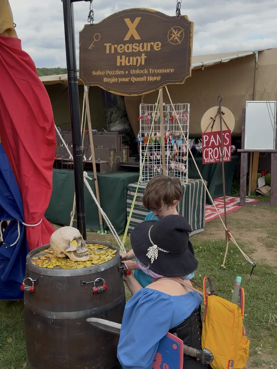 A player in a blue gown and a young adventurer, seen from behind, working the padlocks on a treasure barrel under the X Treasure Hunt sign