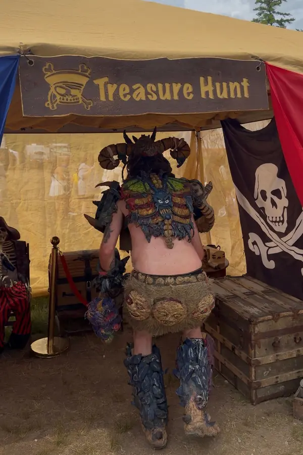 A player in a horned creature costume, seen from behind, at the X Treasure Hunt tent beside the treasure chests and the Jolly Roger