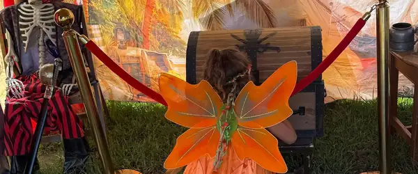 A young adventurer in orange fairy wings, seen from behind, opening the treasure chest in the X Treasure Hunt tent